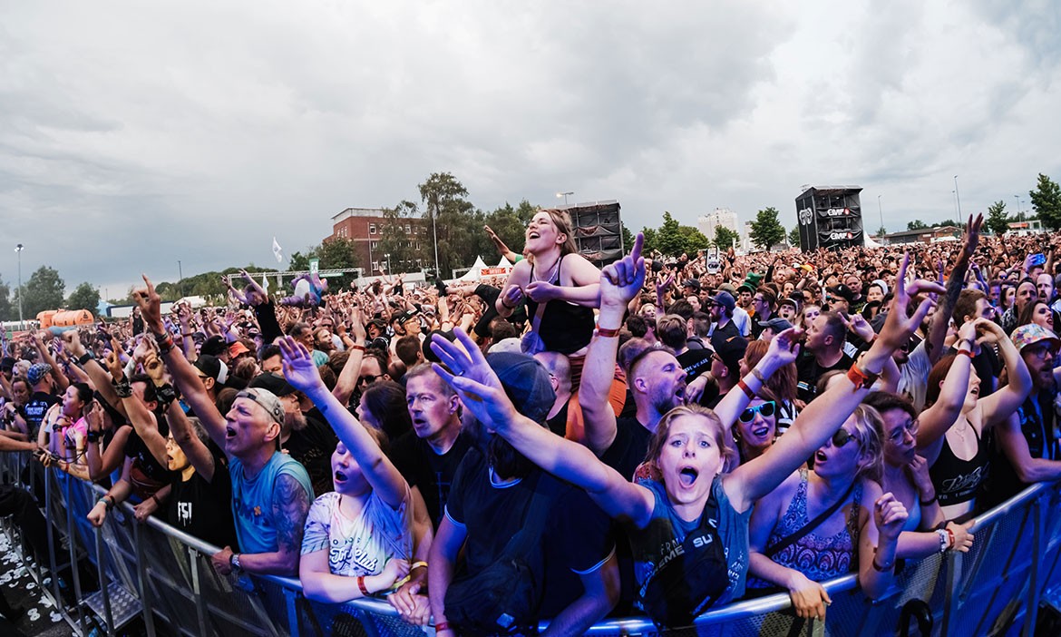 Große Menschenmenge auf dem Vainstream Rockfest Münster, dicht gedrängt vor der Bühne. Viele Besucher und Besucherinnen strecken die Arme in die Luft, jubeln oder singen mit. In der Mitte sitzt eine Frau auf den Schultern einer anderen Person und feiert ausgelassen. Der Himmel ist bewölkt, das Licht auf der Menge ist teils in bläuliche Töne getaucht. Im Hintergrund sind Lautsprechertürme, Festivalzelte und einige Bäume zu erkennen. Die Stimmung ist energiegeladen und euphorisch.