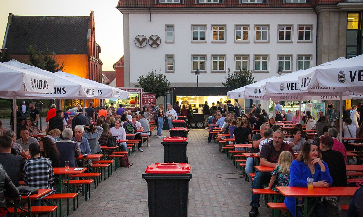 Menschen sitzen an langen Bierzeltgarnituren unter weißen Veltins-Sonnenschirmen auf einem belebten Platz bei Abenddämmerung. Im Hintergrund ist ein Gebäude mit Imbissständen zu sehen, in der Mitte stehen Mülltonnen. Die Stimmung ist gesellig und entspannt.