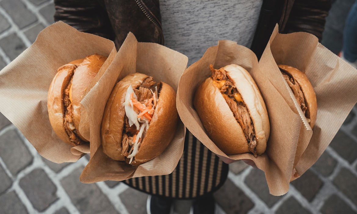 Eine Person in Lederjacke hält vier in braunes Papier gewickelt Burger in die Kamera.