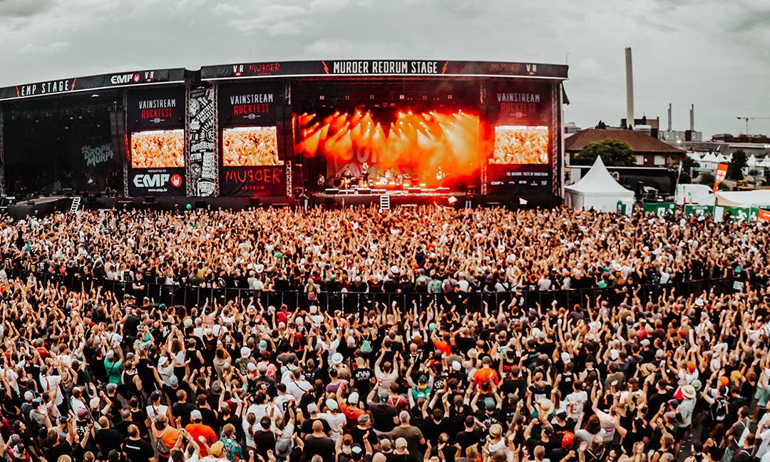 Große Menschenmenge vor einer Open-Air-Bühne beim Vainstream Rockfest in Münster. Die Bühne ist mit roten Lichtern beleuchtet, auf mehreren Screens ist der Schriftzug „Vainstream Rockfest“ zu sehen. Die Menge reckt die Hände in die Höhe, der Himmel ist bewölkt und grau. Im Hintergrund sind Industriegebäude und Festivalstände zu erkennen. Die Stimmung ist ausgelassen und energiegeladen.