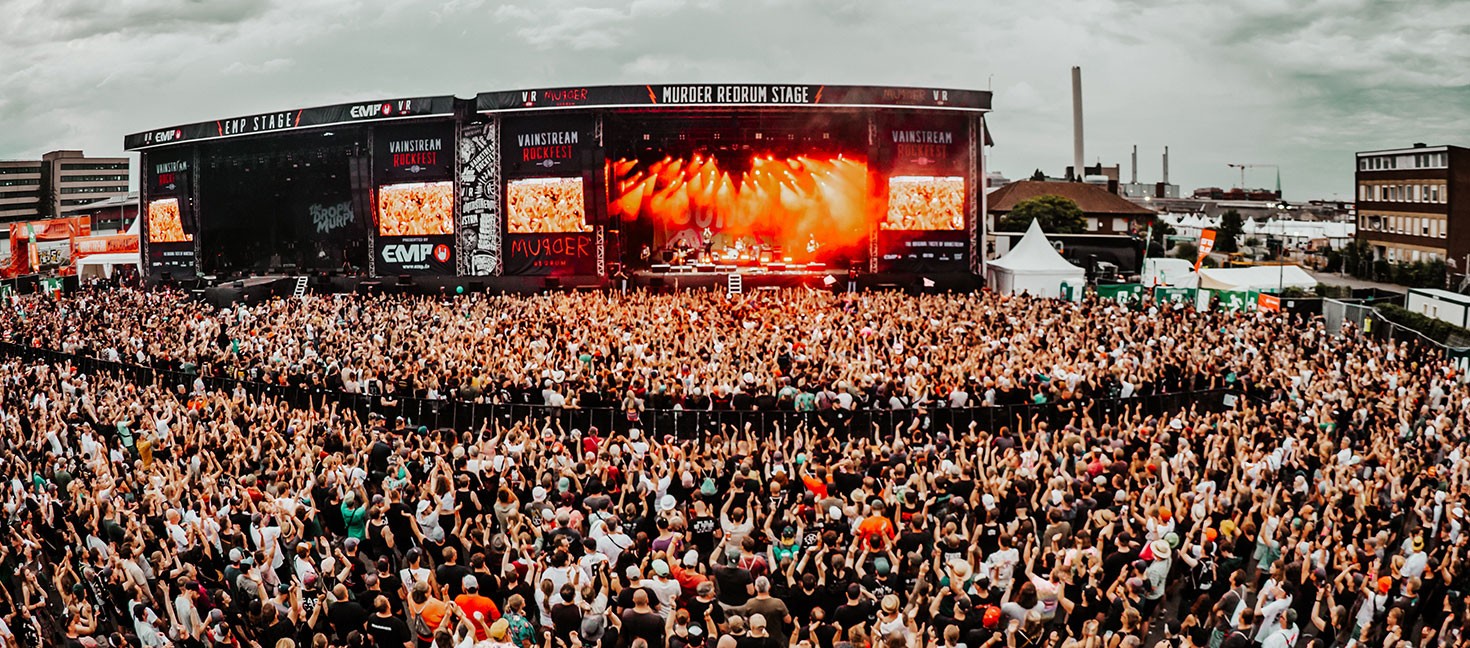Große Menschenmenge vor einer Open-Air-Bühne beim Vainstream Rockfest in Münster. Die Bühne ist mit roten Lichtern beleuchtet, auf mehreren Screens ist der Schriftzug „Vainstream Rockfest“ zu sehen. Die Menge reckt die Hände in die Höhe, der Himmel ist bewölkt und grau. Im Hintergrund sind Industriegebäude und Festivalstände zu erkennen. Die Stimmung ist ausgelassen und energiegeladen.