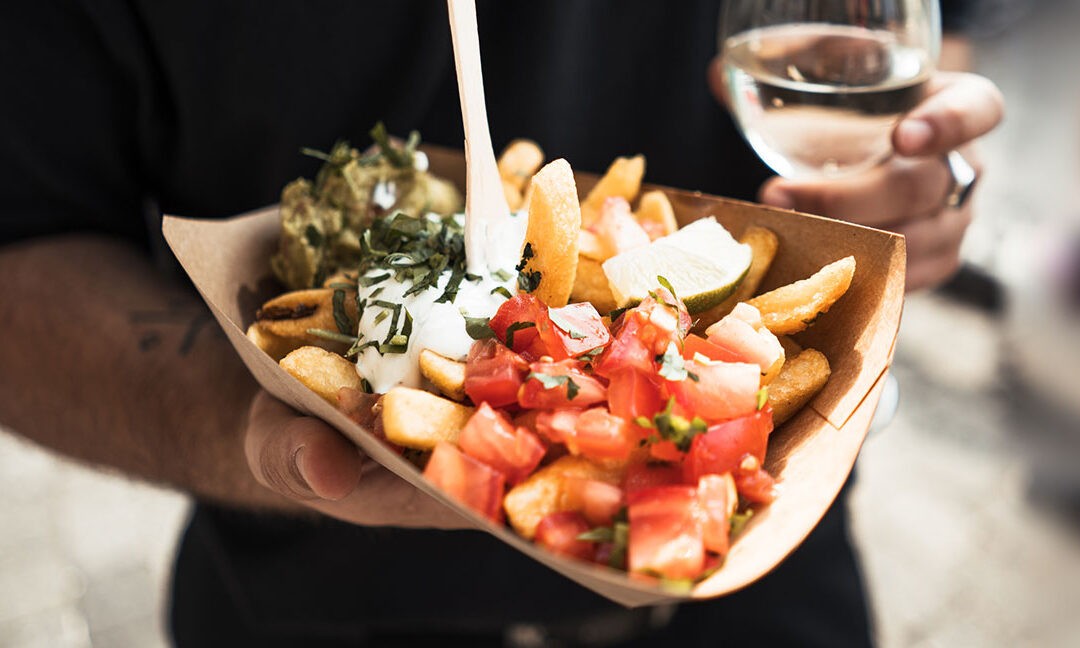 Eine Person hält eine Portion Street Food in einer braunen Pappschale: Pommes mit frischen Tomatenwürfeln, Guacamole, Sour Cream und Kräutern, dazu ein Holzspieß. In der anderen Hand ein Glas Weißwein.