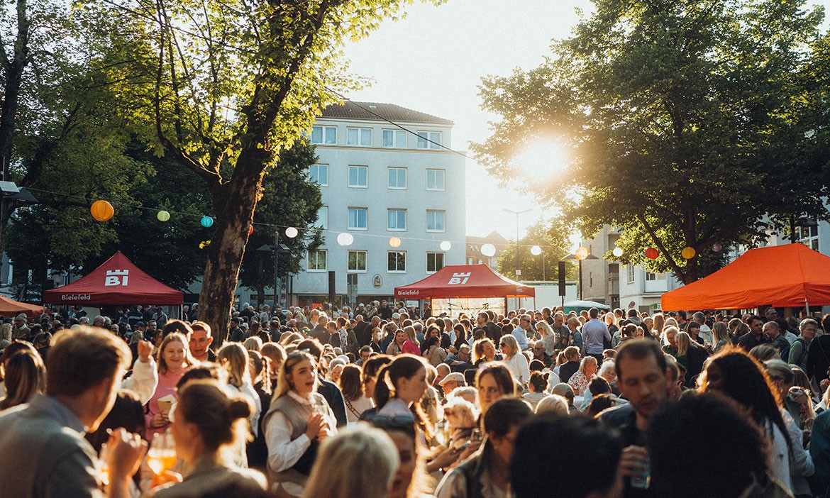 Menschenmenge beim Abendmarkt in Bielefeld unter bunten Lampions, die tiefstehende Sonne sorgt für stimmungsvolles Gegenlicht und sommerliche Atmosphäre zwischen Marktständen und Altbaufassaden.