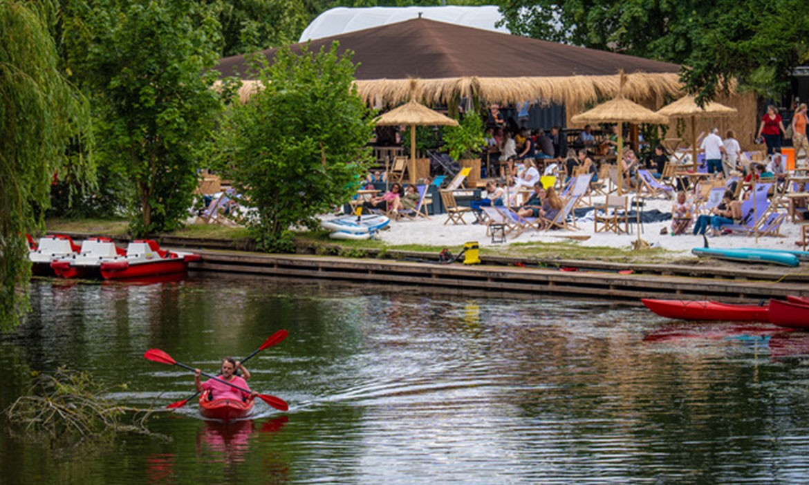 Blick auf die Grinsekatz Braunschweig vom Wasser aus: Im Vordergrund paddelt eine Frau in einem roten Kajak auf der Oker. Am gegenüberliegenden Ufer sitzen zahlreiche Gäste im Sand auf Liegestühlen unter Strohdächern, umgeben von Palmen und Bäumen. Die überdachte Beachbar ist gut besucht, im Hintergrund Tretboote und weitere Wasserfahrzeuge – lebendige Sommerstimmung am Stadtstrand im Grünen.