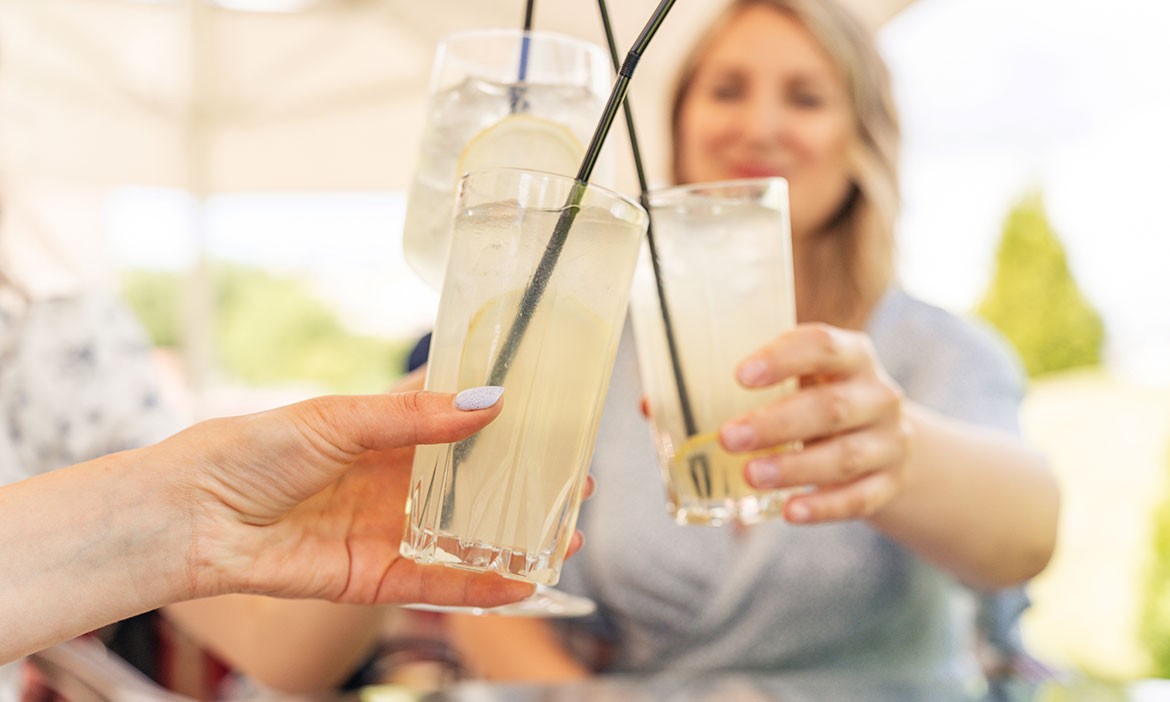 Drei Personen stoßen im Freien mit Longdrinkgläsern voller Eis, Zitronenscheiben und Strohhalmen an. Im Hintergrund sitzt eine lächelnde Frau unscharf im Bild, sonnige Sommeratmosphäre.