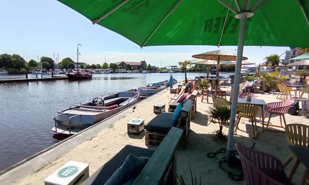 Strandbereich am Wasser mit Sandboden, bunten Stühlen, Sonnenschirmen und Palmen beim Delftstrand Emden. Im Vordergrund Sitzgelegenheiten unter einem großen grünen Sonnenschirm, im Hintergrund liegen Boote am Kai, dahinter sind Yachten und Hafengebäude zu sehen – entspannte Sommeratmosphäre direkt am Wasser.