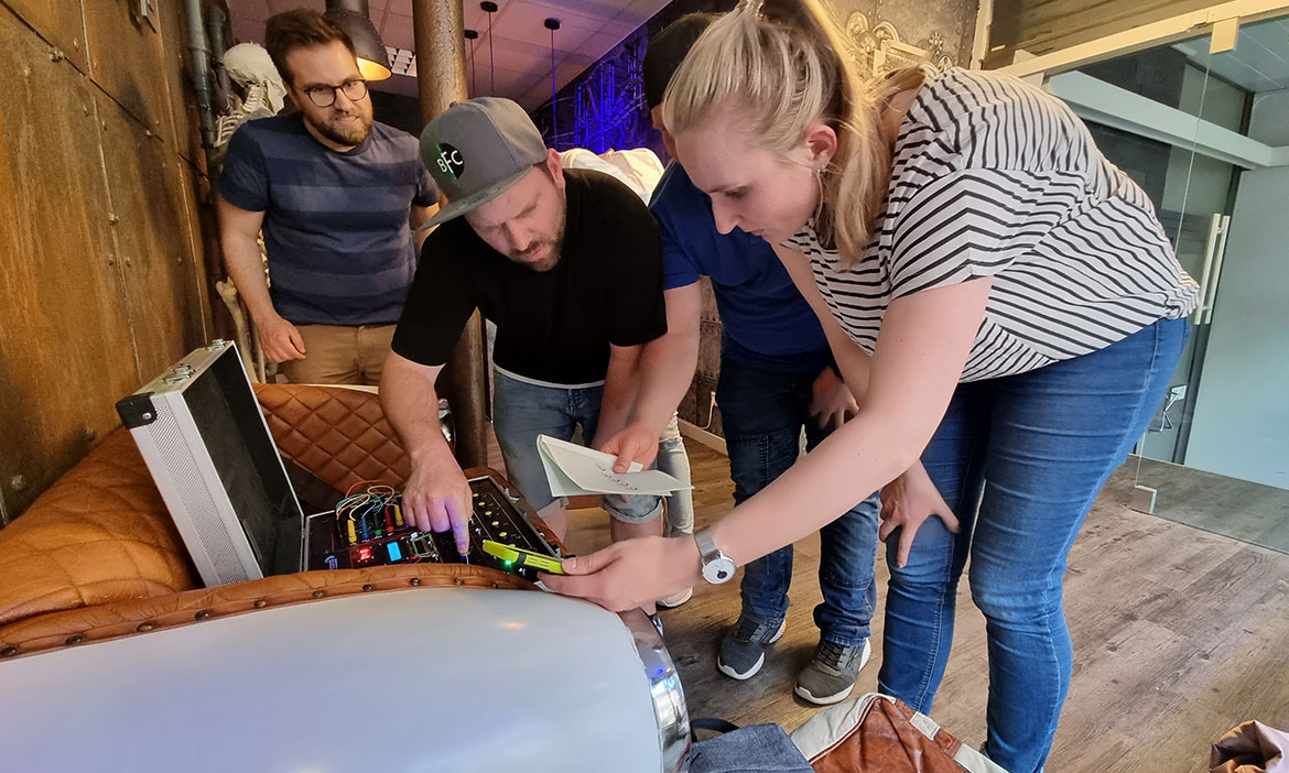 Vier Spieler des Outdoor Escape Games „Mission: Bombe“ vom try2escape in Rheine schauen sich gemeinsam einen präparierten Rätselkoffer an. Sie befinden sich in einem der Escape Rooms. Eine Spielerin durchsucht konzentriert das Innenleben des Koffers mit farbigen Kabeln, während ihre Teamkollegen Hinweise analysieren. 