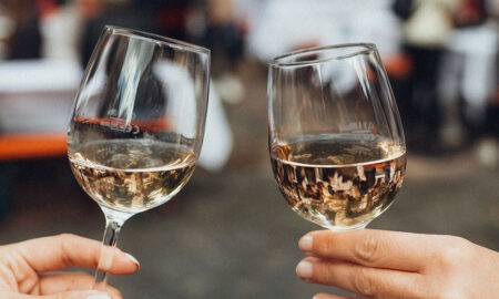 Zwei Hände stoßen mit gefüllten Weißweingläsern an, im Hintergrund verschwommen eine gesellige Szenerie mit Bierbänken und Menschen auf dem Abendmarkt in Bielefeld.