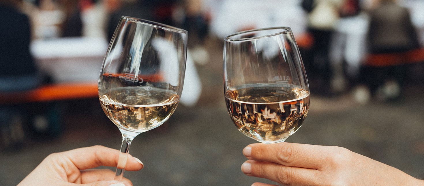 Zwei Hände stoßen mit gefüllten Weißweingläsern an, im Hintergrund verschwommen eine gesellige Szenerie mit Bierbänken und Menschen auf dem Abendmarkt in Bielefeld.