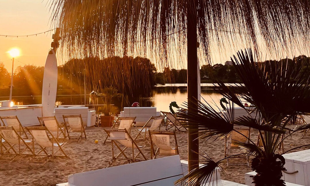 Strandbereich mit Liegestühlen und Palmen bei Sonnenuntergang, Blick auf einen See – entspannte Atmosphäre in einem Beachclub in NRW oder Niedersachsen