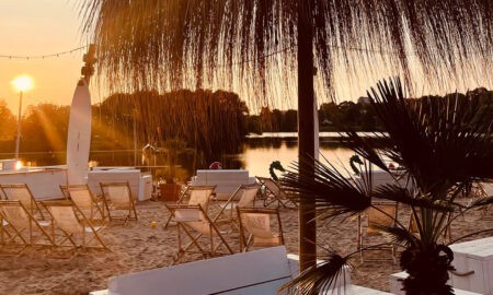Strandbereich mit Liegestühlen und Palmen bei Sonnenuntergang, Blick auf einen See – entspannte Atmosphäre in einem Beachclub in NRW oder Niedersachsen
