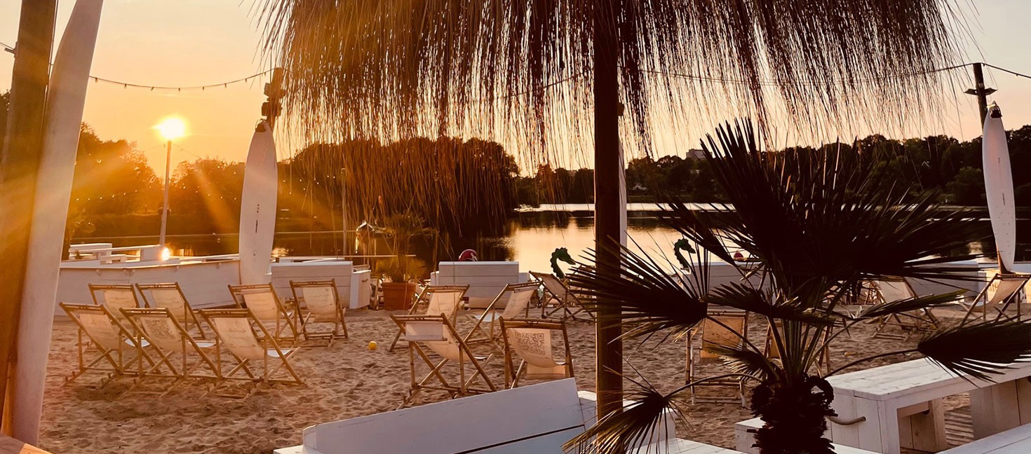 Strandbereich mit Liegestühlen und Palmen bei Sonnenuntergang, Blick auf einen See – entspannte Atmosphäre in einem Beachclub in NRW oder Niedersachsen