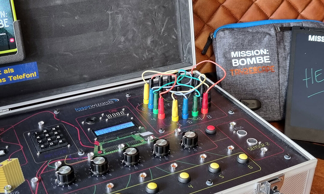 Ein mobiles Escape-Game-Set mit dem Titel „MISSION: BOMBE“. Zu sehen ist ein silberner Koffer mit technischen Bedienelementen, Kabeln, bunten Steckverbindungen und Drehknöpfen. Auf der Innenseite des Kofferdeckels ist ein Smartphone befestigt mit der Aufschrift „Entsperrt als erstes das Telefon!“. Daneben liegen ein Tablet mit der Aufschrift „HELP!“ sowie eine Tasche mit dem Logo „MISSION: BOMBE – Das Escape Game“.
