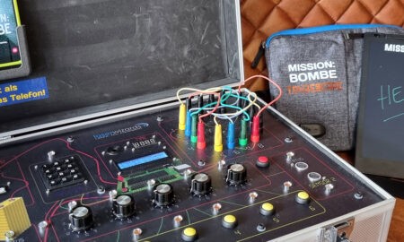 Ein mobiles Escape-Game-Set mit dem Titel „MISSION: BOMBE“. Zu sehen ist ein silberner Koffer mit technischen Bedienelementen, Kabeln, bunten Steckverbindungen und Drehknöpfen. Auf der Innenseite des Kofferdeckels ist ein Smartphone befestigt mit der Aufschrift „Entsperrt als erstes das Telefon!“. Daneben liegen ein Tablet mit der Aufschrift „HELP!“ sowie eine Tasche mit dem Logo „MISSION: BOMBE – Das Escape Game“.