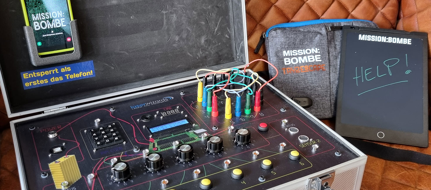 Ein mobiles Escape-Game-Set mit dem Titel „MISSION: BOMBE“. Zu sehen ist ein silberner Koffer mit technischen Bedienelementen, Kabeln, bunten Steckverbindungen und Drehknöpfen. Auf der Innenseite des Kofferdeckels ist ein Smartphone befestigt mit der Aufschrift „Entsperrt als erstes das Telefon!“. Daneben liegen ein Tablet mit der Aufschrift „HELP!“ sowie eine Tasche mit dem Logo „MISSION: BOMBE – Das Escape Game“.