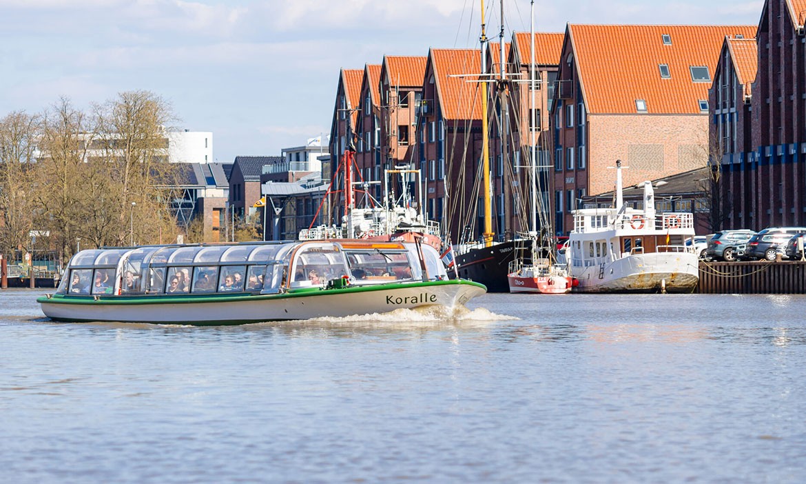 Ausflugsschiff ‚Koralle‘ mit Glasdach fährt durch den Hafen von Leer, im Hintergrund historische Speicherhäuser und weitere Schiffe.