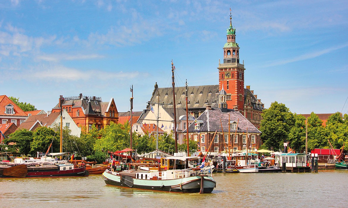 Blick auf den Hafen von Leer mit historischen Segelschiffen im Vordergrund und dem prächtigen Rathaus im Hintergrund bei sonnigem Wetter.