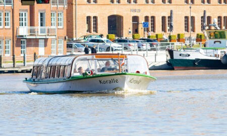 Das Ausflugsschiff ‚Koralle‘ fährt durch den Hafen von Leer, im Hintergrund historische Backsteingebäude und weitere festgemachte Schiffe.