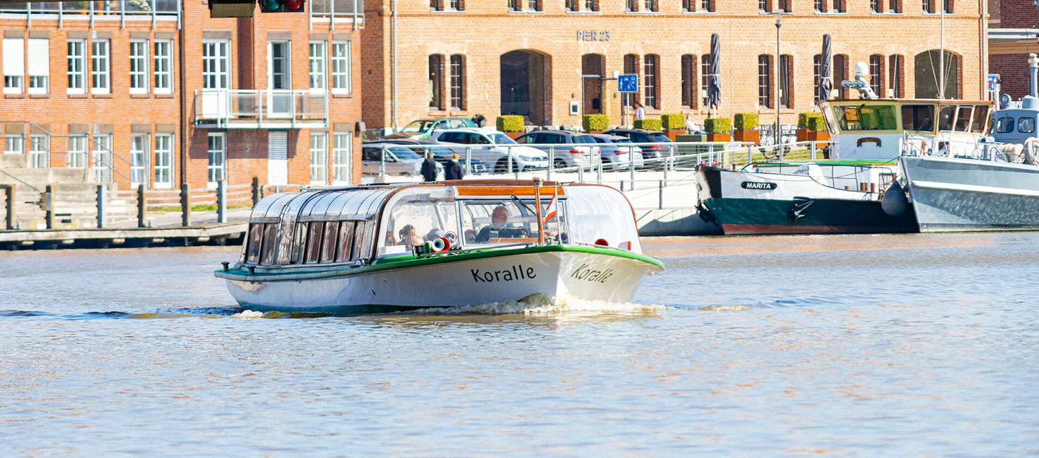 Das Ausflugsschiff ‚Koralle‘ fährt durch den Hafen von Leer, im Hintergrund historische Backsteingebäude und weitere festgemachte Schiffe.