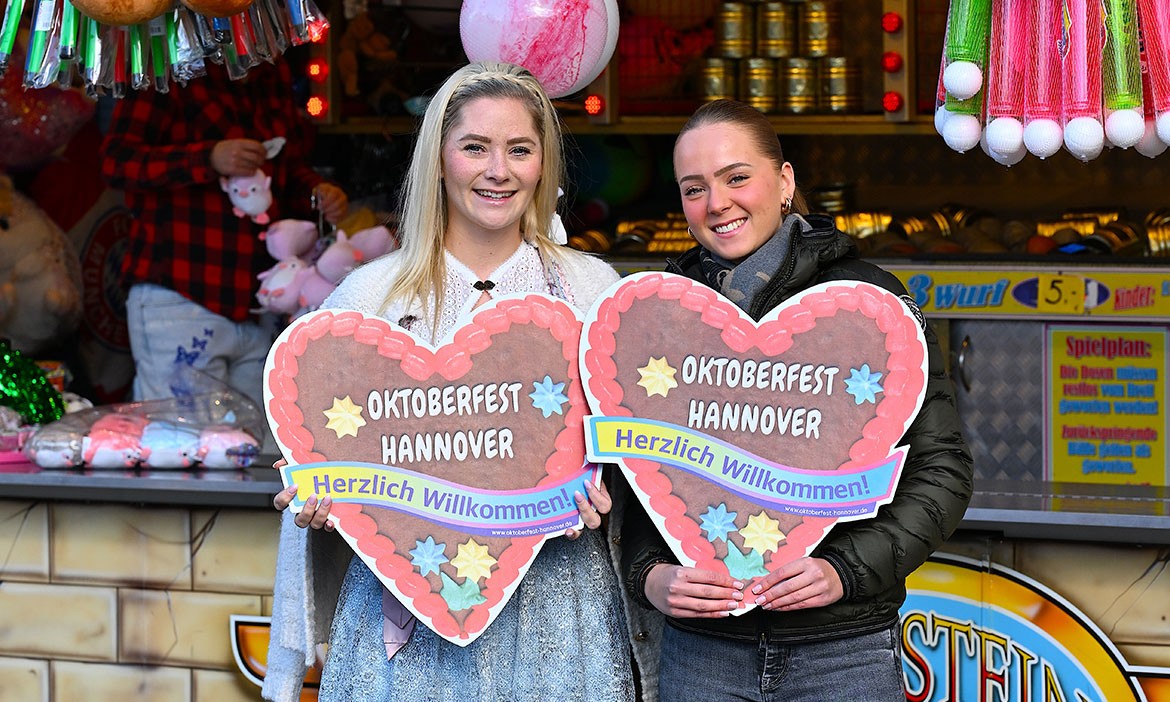 Zwei lächelnde Frauen stehen auf dem Oktoberfest in Hannover vor einem Schaustellerstand und halten jeweils ein großes Lebkuchenherz mit der Aufschrift „Oktoberfest Hannover – Herzlich Willkommen!“ in den Händen.