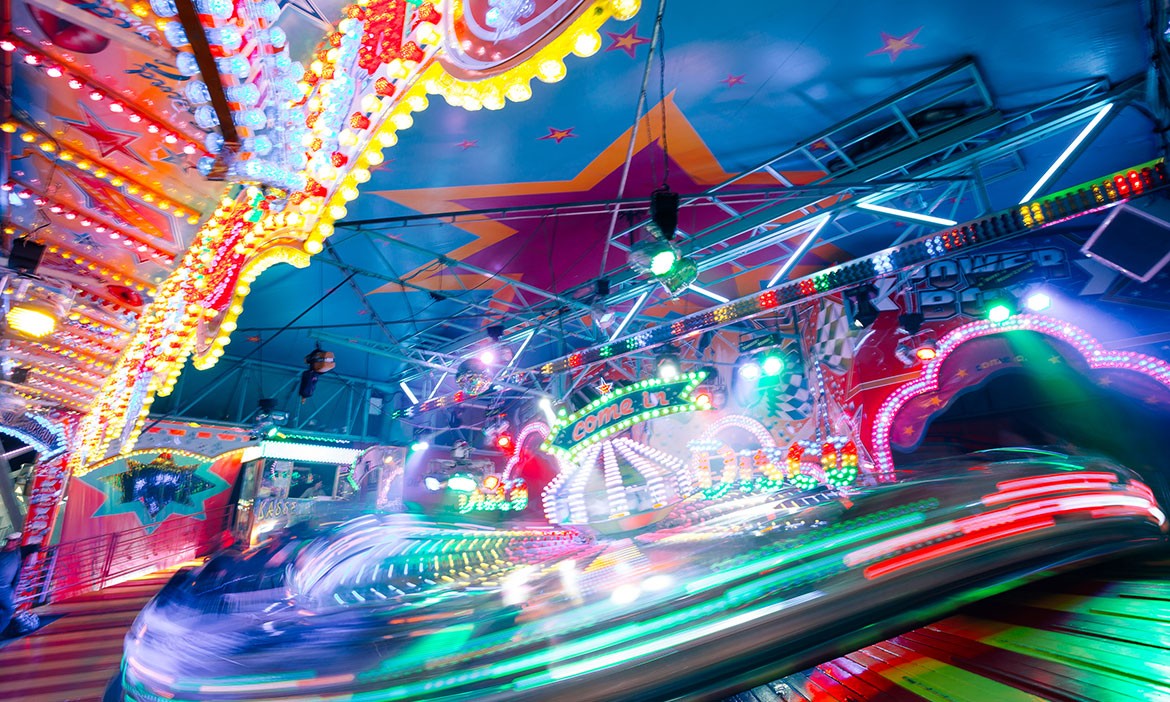 Das Bild zeigt ein buntes Fahrgeschäft auf einem Rummelplatz, beleuchtet von zahlreichen Lichtern in verschiedenen Farben, darunter Rot, Gelb, Blau und Grün. Die Decke des Fahrgeschäfts ist mit großen, farbenfrohen Sternen und Lichtelementen dekoriert, die eine festliche Atmosphäre schaffen. Die Bewegungsunschärfe der sich drehenden Fahrkabinen verleiht dem Bild eine dynamische Note. Im Hintergrund sind leuchtende Schilder und Dekorationen zu sehen, die den typischen Jahrmarkt-Flair verstärken. Die fröhliche, lebendige Szenerie fängt die Energie und den Spaß eines Rummelplatzes ein.