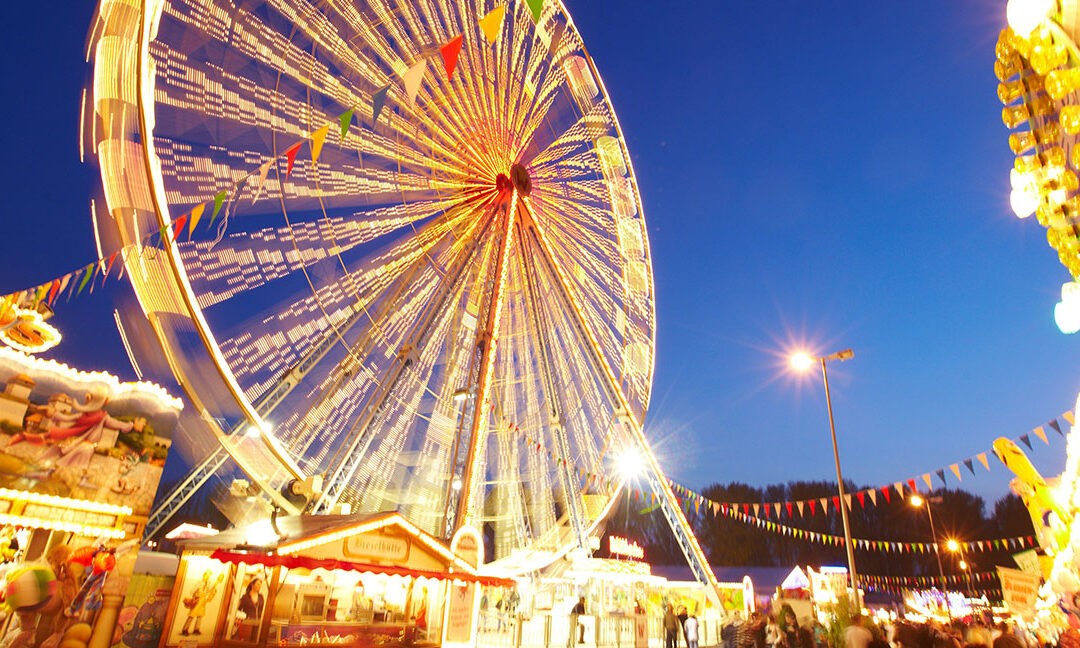 Das Bild zeigt eine fröhliche Jahrmarkt-Szene bei Nacht mit einem großen, beleuchteten Riesenrad im Zentrum, das in gelben und weißen Lichtern erstrahlt. Bunte Wimpelketten hängen quer durch das Bild und verstärken die festliche Atmosphäre. Im Vordergrund sind beleuchtete Verkaufsstände mit farbenfrohen Schildern und Dekorationen zu sehen, die typische Jahrmarkt-Attraktionen wie Spiele und Snacks anbieten. Der Himmel ist tiefblau, was die Lichter des Riesenrads und der Stände noch intensiver leuchten lässt. Die Szenerie ist belebt, mit Besuchern, die den Rummelplatz genießen.