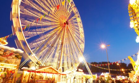 Das Bild zeigt eine fröhliche Jahrmarkt-Szene bei Nacht mit einem großen, beleuchteten Riesenrad im Zentrum, das in gelben und weißen Lichtern erstrahlt. Bunte Wimpelketten hängen quer durch das Bild und verstärken die festliche Atmosphäre. Im Vordergrund sind beleuchtete Verkaufsstände mit farbenfrohen Schildern und Dekorationen zu sehen, die typische Jahrmarkt-Attraktionen wie Spiele und Snacks anbieten. Der Himmel ist tiefblau, was die Lichter des Riesenrads und der Stände noch intensiver leuchten lässt. Die Szenerie ist belebt, mit Besuchern, die den Rummelplatz genießen.