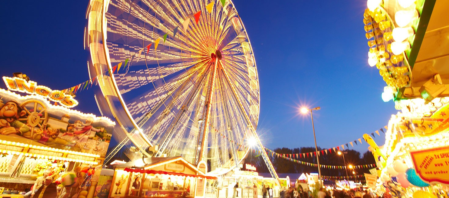 Das Bild zeigt eine fröhliche Jahrmarkt-Szene bei Nacht mit einem großen, beleuchteten Riesenrad im Zentrum, das in gelben und weißen Lichtern erstrahlt. Bunte Wimpelketten hängen quer durch das Bild und verstärken die festliche Atmosphäre. Im Vordergrund sind beleuchtete Verkaufsstände mit farbenfrohen Schildern und Dekorationen zu sehen, die typische Jahrmarkt-Attraktionen wie Spiele und Snacks anbieten. Der Himmel ist tiefblau, was die Lichter des Riesenrads und der Stände noch intensiver leuchten lässt. Die Szenerie ist belebt, mit Besuchern, die den Rummelplatz genießen.