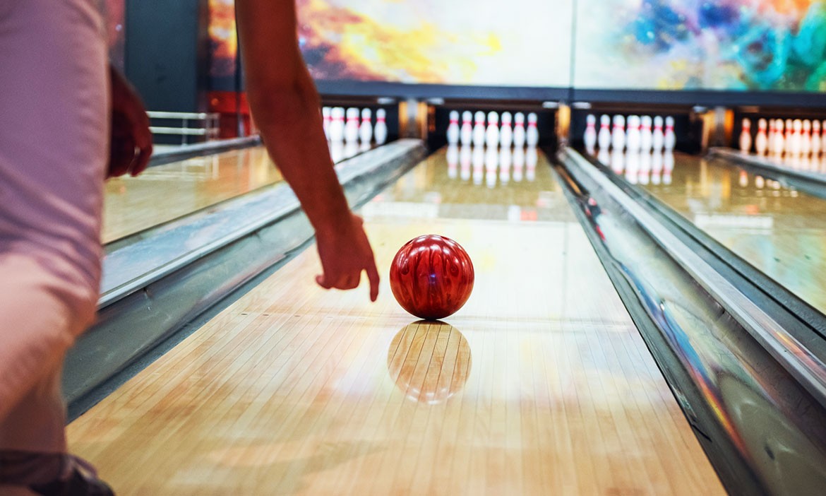 Eine rote Bowlingkugel rollt auf einer glänzenden Bahn in Richtung Pins. Im Vordergrund ist der Arm einer Person beim Wurf zu sehen. Die Szene ist dynamisch und zeigt den Moment kurz nach dem Abwurf.