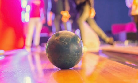 Bowlingkugel im Fokus auf der glänzenden Bahn, im Hintergrund unscharf eine Gruppe junger Menschen beim Spielen. Farbige Lichter sorgen für lebendige, dynamische Stimmung.