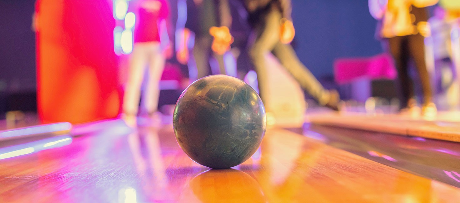 Bowlingkugel im Fokus auf der glänzenden Bahn, im Hintergrund unscharf eine Gruppe junger Menschen beim Spielen. Farbige Lichter sorgen für lebendige, dynamische Stimmung.