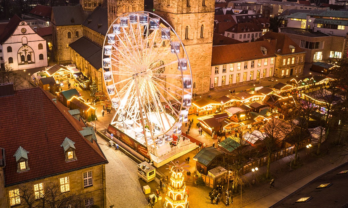 Luftaufnahme des festlich beleuchteten Weihnachtsmarkts in Osnabrück bei Abenddämmerung, mit einem großen Riesenrad auf dem Marktplatz, umgeben von Buden, Lichtern und historischen Gebäuden.