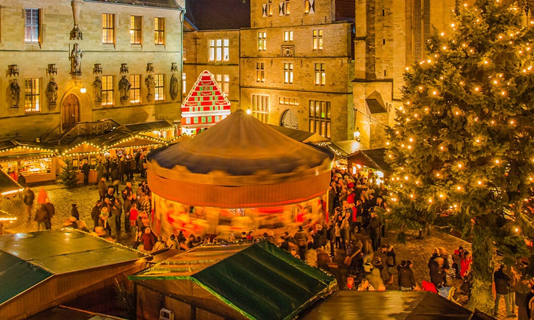 Blick auf den festlich beleuchteten Weihnachtsmarkt in Osnabrück mit großem Weihnachtsbaum, Karussell und vielen Besucherinnen und Besuchern vor dem historischen Rathaus.