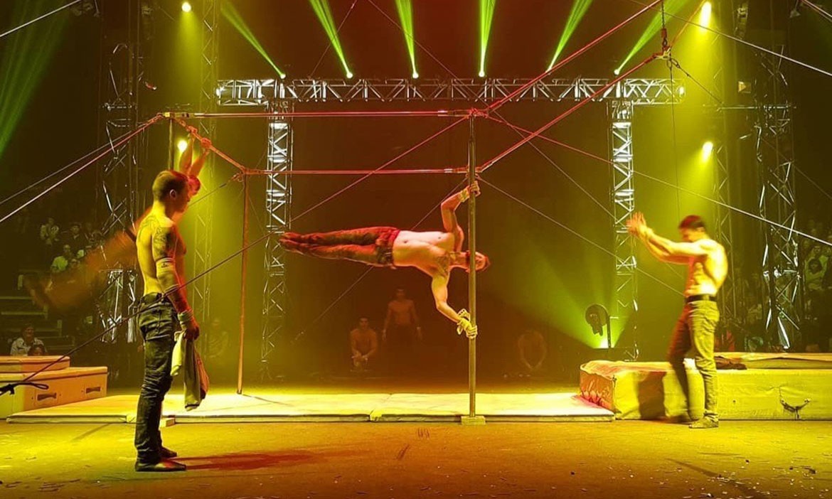 Drei männliche Artisten stehen auf der Bühne der Flic Flac Xmas Show in Bielefeld. In der Mitte hält einer von ihnen eine anspruchsvolle Pose an einer senkrechten Stange. Die Szene ist in gelb-grünes Licht getaucht, umgeben von Drahtseilkonstruktionen und Showbeleuchtung. Im Hintergrund sind Zuschauer erkennbar.