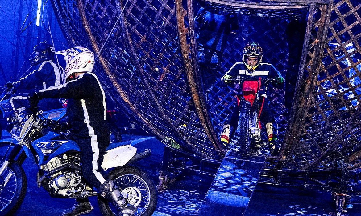 Das Bild zeigt mehrere Motorradfahrer in schwarzer Showkleidung mit Helmen, die sich in und um die sogenannte „Todeskugel“ im Zelt von Flic Flac bewegen. Ein Fahrer verlässt gerade die große, kugelförmige Stahlkonstruktion über eine Rampe. Die Szene ist in blaues Showlicht getaucht und vermittelt den Eindruck einer dynamischen, technisch anspruchsvollen Stuntperformance während der Flic Flac Xmas Show.