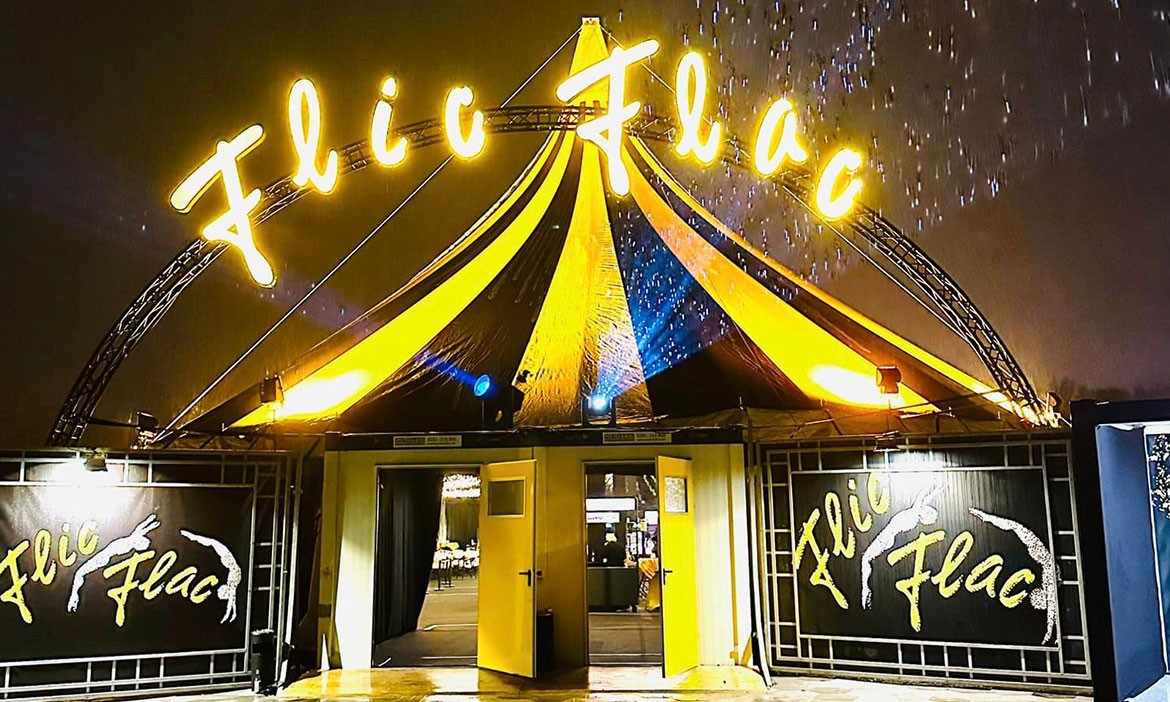 Das Bild zeigt den Eingang des Zirkuszelts von Flic Flac auf dem Festplatz an der Radrennbahn in Bielefeld. Das Zelt ist in den typischen Farben Schwarz und Gelb gehalten. Über dem Eingang leuchten die Worte „Flic Flac“ in großen, hellen Buchstaben. Es ist Nacht, und der regennasse Boden spiegelt das Licht des Zelts. Die Türen stehen offen und geben den Blick auf das beleuchtete Vorzelt frei.