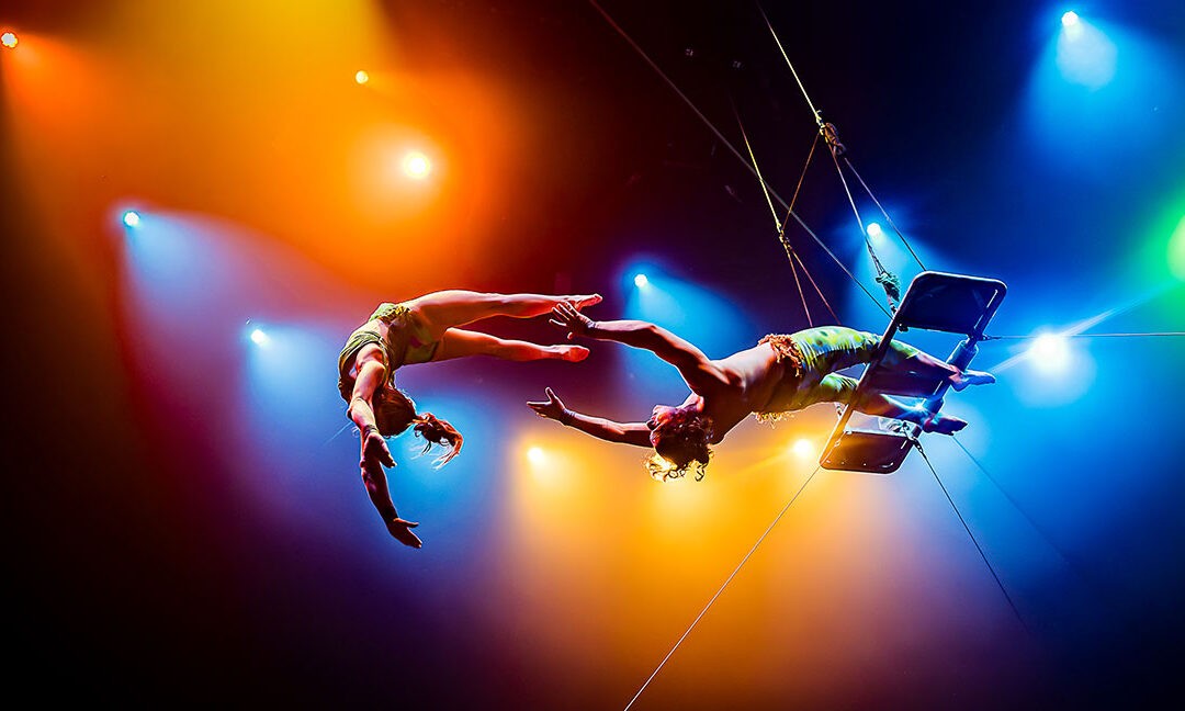 Zwei Artist:innen führen einen Trapezsprung unter farbigem Bühnenlicht aus. Eine Person fliegt frei durch die Luft, die andere hängt kopfüber am Trapez. In der Mitte des Raums greifen sie sich an den Händen. Das warme Orange- und kühle Blaulicht verstärkt die Dynamik und Spannung der Szene.