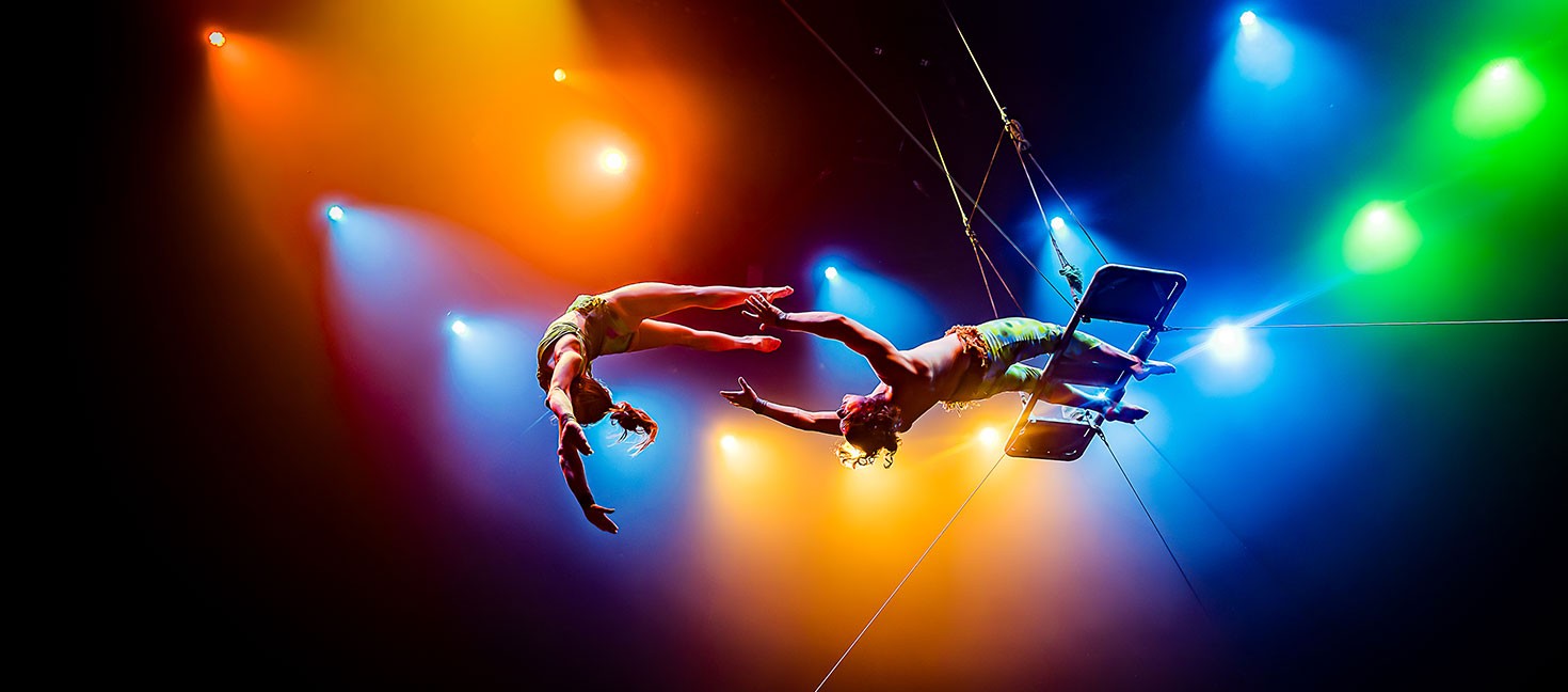 Zwei Artist:innen führen einen Trapezsprung unter farbigem Bühnenlicht aus. Eine Person fliegt frei durch die Luft, die andere hängt kopfüber am Trapez. In der Mitte des Raums greifen sie sich an den Händen. Das warme Orange- und kühle Blaulicht verstärkt die Dynamik und Spannung der Szene.