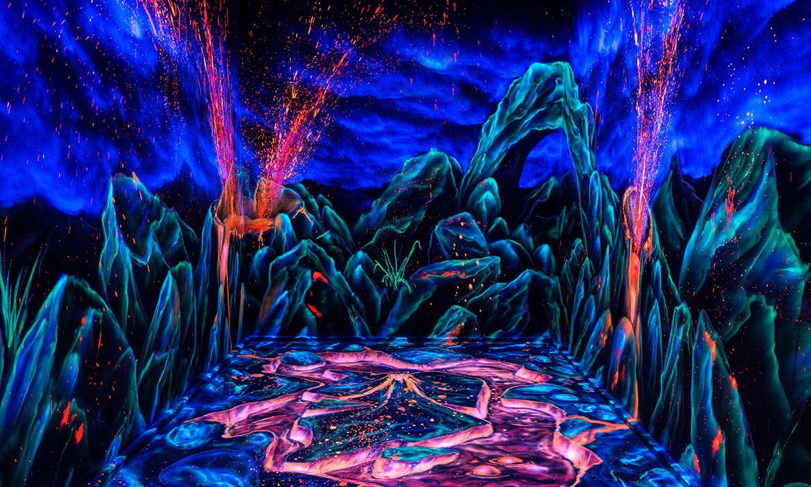 Ein komplett in Schwarzlicht gestalteter Raum zeigt eine spektakuläre Fantasie-Felsenlandschaft in Blau- und Grüntönen. Orange-rote „Funken“ und lavaartige Farbakzente leuchten an den Wänden, während sich in der Mitte eine bunt schimmernde Bahn durch die Szene zieht.