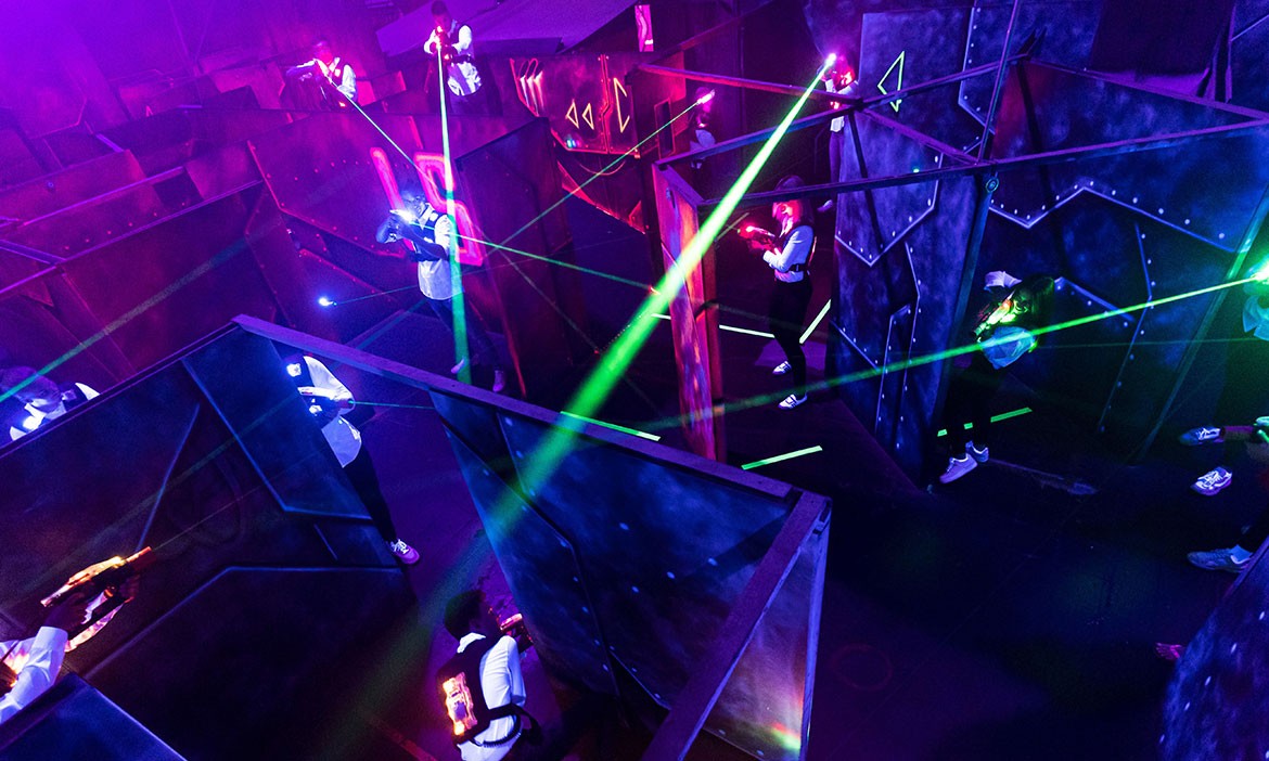 In einer dunklen, futuristisch wirkenden Laser-Tag-Arena stehen mehrere Spielerinnen und Spieler in den Gängen und zielen mit ihren Markierern. Grüne Laserstrahlen schneiden durch den Raum, während Neonmarkierungen und violettes Licht die verwinkelten Wände und Ecken der Arena betonen.