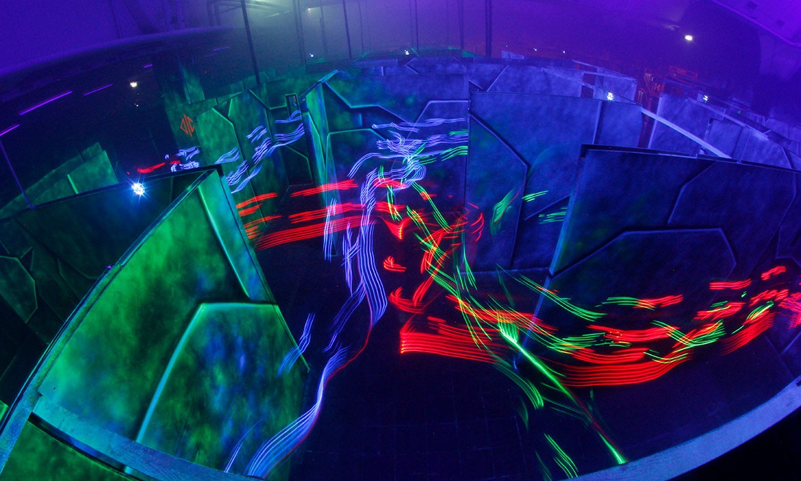 Eine Aufnahme von oben zeigt ein verwinkeltes Arenalabyrinth mit hohen Wänden in dunklen Blau- und Grüntönen. Durch Langzeitbelichtung ziehen sich rote, grüne und blaue Lichtspuren durch die Gänge – sie vermitteln Tempo, Action und den typischen Neon-Look einer Laser-Tag-Umgebung.