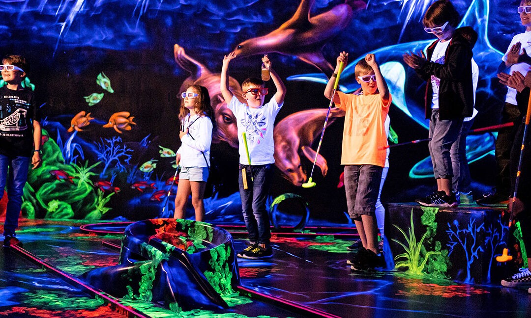 Eine Gruppe aus Kindern und Erwachsenen spielt in einer Schwarzlicht-Anlage Minigolf. Alle tragen 3D-Brillen, halten bunte Schläger in der Hand und jubeln vor einer neonfarbenen Unterwasser-Wandmalerei mit Haien, Fischen und Korallen.