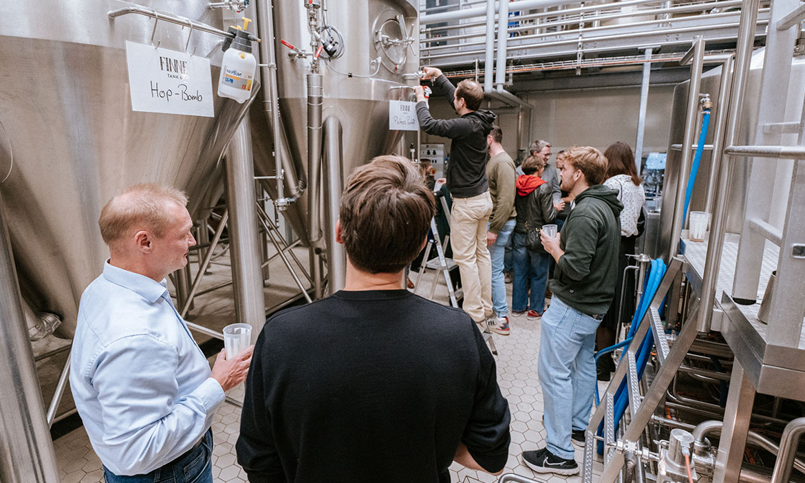 Gruppe von Personen während einer Brauereiführung in einer Produktionshalle mit großen Edelstahltanks; eine Person steht auf einer Leiter und befestigt ein Schild mit der Aufschrift „FINNE Tank 6 Hop Bomb“ an einem Tank, während andere mit Probiergläsern danebenstehen.