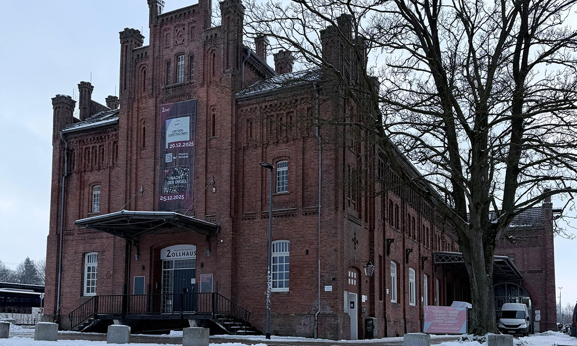 Schräg von vorn fotografiertes Backsteingebäude des Zollhaus Leer im Winter. Vor dem Haupteingang führt eine Treppe zu einer überdachten Rampe, darüber hängt ein großes Veranstaltungsbanner an der Fassade. Auf dem Boden liegt Schnee, die Bäume sind kahl und der Himmel ist grau – die Szene wirkt kalt und ruhig.