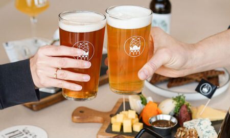 Zwei Personen stoßen mit hellen, bernsteinfarbenen Biergläsern mit Schaumkrone an. Im Hintergrund stehen Käse- und Snackbrett sowie weitere Biere auf einem Tisch.