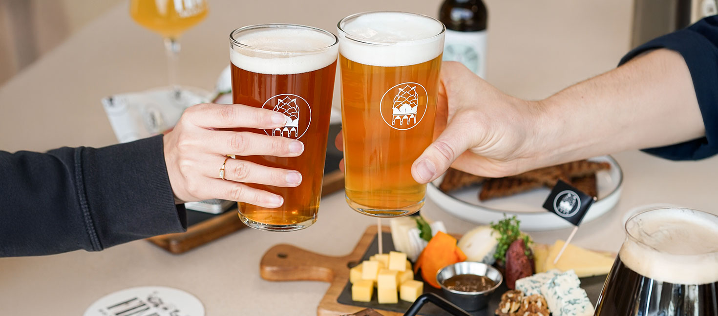 Zwei Personen stoßen mit hellen, bernsteinfarbenen Biergläsern mit Schaumkrone an. Im Hintergrund stehen Käse- und Snackbrett sowie weitere Biere auf einem Tisch.
