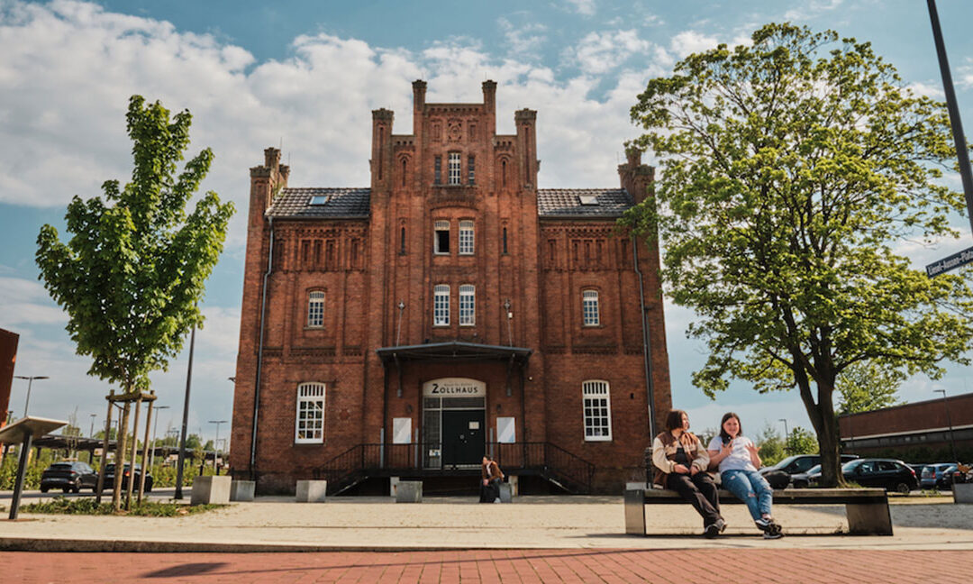 Frontalaufnahme des Zollhaus Leer an einem sonnigen Tag. Das große Backsteingebäude steht mittig im Bild, davor ein weiter Platz mit Pflaster und Sitzgelegenheiten. Rechts und links rahmen grüne Bäume die Szene, im Hintergrund sind parkende Autos zu sehen. Zwei Personen sitzen entspannt auf einer Bank im Vordergrund, während die Fassade des Zollhauses klar und warm im Tageslicht wirkt.
