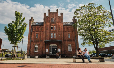 Frontalaufnahme des Zollhaus Leer an einem sonnigen Tag. Das große Backsteingebäude steht mittig im Bild, davor ein weiter Platz mit Pflaster und Sitzgelegenheiten. Rechts und links rahmen grüne Bäume die Szene, im Hintergrund sind parkende Autos zu sehen. Zwei Personen sitzen entspannt auf einer Bank im Vordergrund, während die Fassade des Zollhauses klar und warm im Tageslicht wirkt.