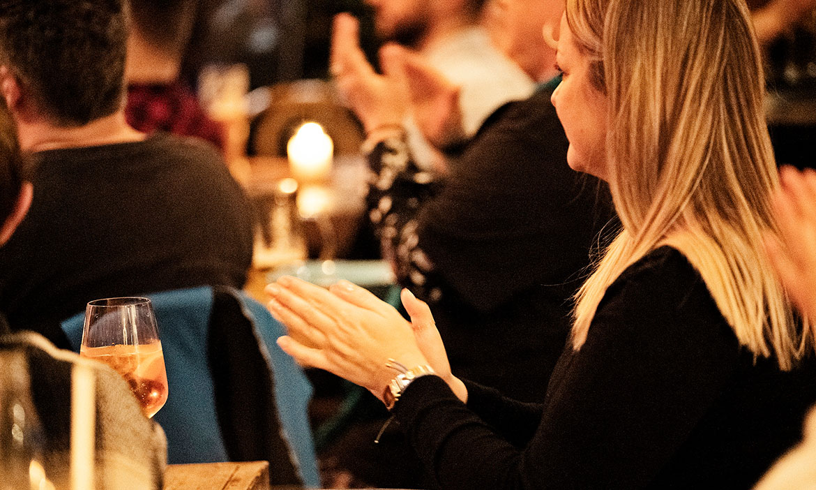 Eine Frau sitzt an einem Tisch mit Getränken und applaudiert während einer Comedy-Veranstaltung. Um sie herum sitzen weitere Gäste in gemütlicher Abendatmosphäre bei Kerzenlicht und verfolgen aufmerksam das Programm.