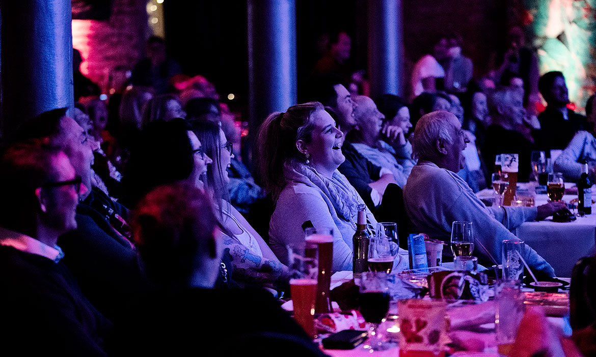 Ein großer Saal voller Gäste sitzt an Tischen mit Getränken und schaut zur Bühne. Viele Menschen lachen und reagieren begeistert auf eine Comedy-Performance, während farbige Bühnenbeleuchtung den Raum in Blau- und Pinktöne taucht.